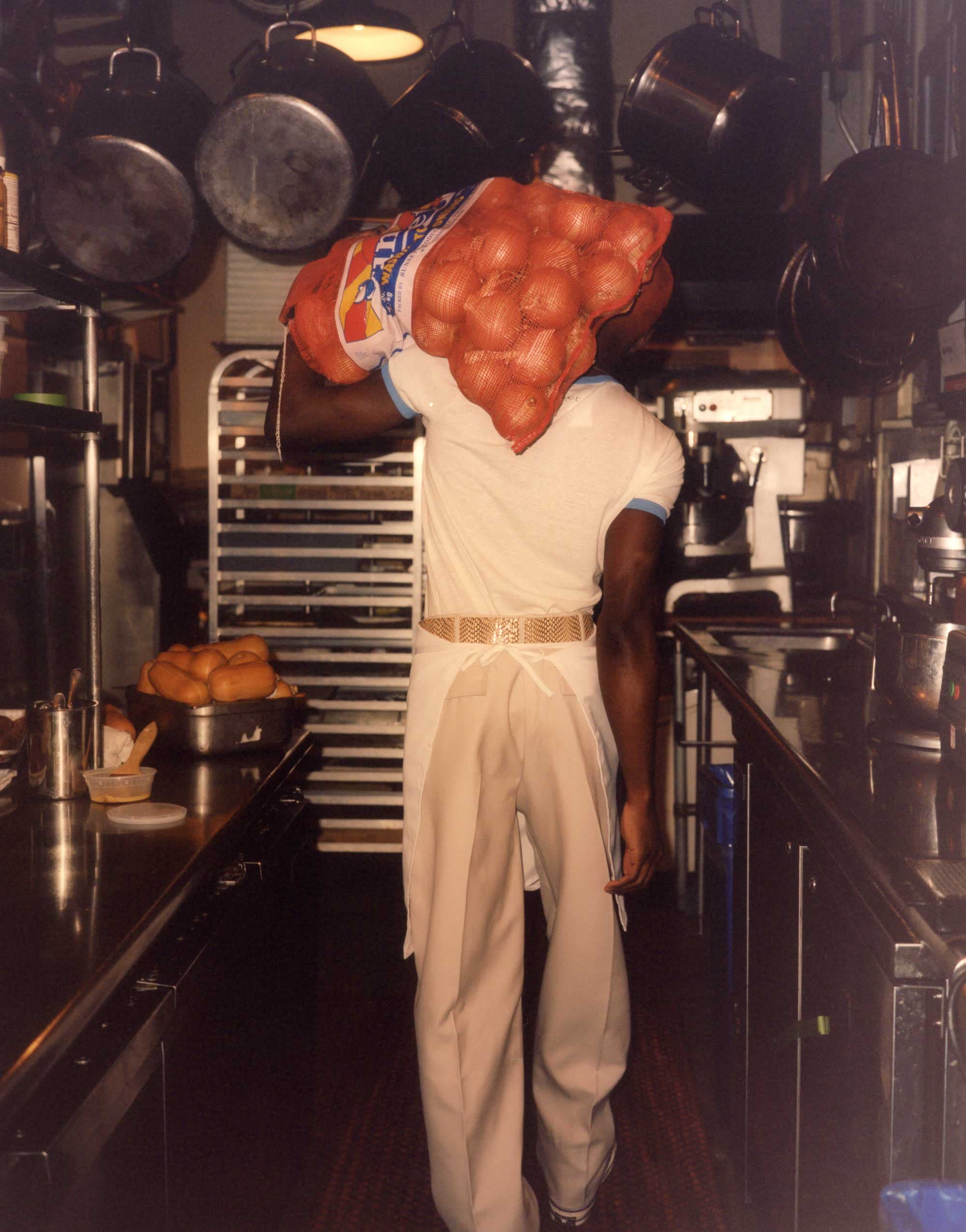 A kitchen porter carrying a sack of onions down the line, past hanging pans